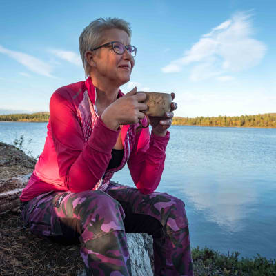Nainen pinkissä takissa istuu kiven päällä kuksa käsissä, hymyilee, taustalla järvi ja erämaata.