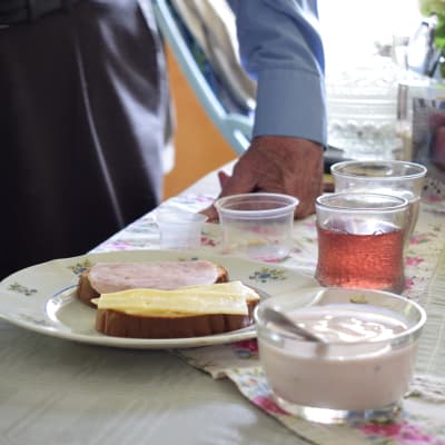 En äldre persons han på ett bord intill en frukost med juice, smörgås och yoghurt,