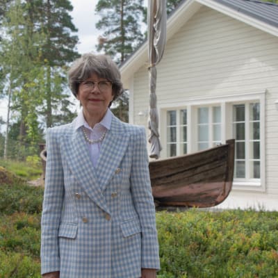 Hanna Kaarina Syrjäläinen vid Villa Albert, i bakgrunden ett vitt hus och en träbåt. 