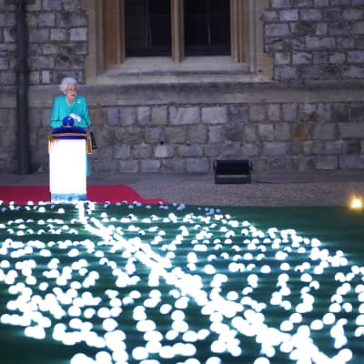 Kuningatar Elisabeth seisoo hehkuvan korokkeen edessä. Korokkeen edestä levittäytyy nurmelle valopallojen ketjuja.