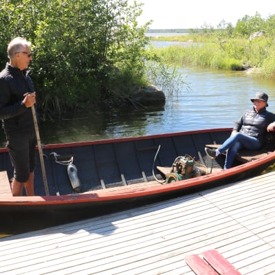 En kvinna sitter i aktern av en gammal puttputt-båt som ligger förtöjd vid en brygga. I fören står en man.