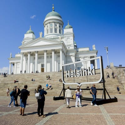 Turister poserar framför en "Helsinki"-skylt på Senatstorget framför Domkyrkans trappor.