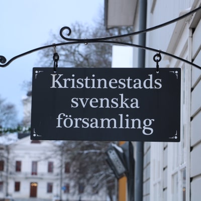 En skylt hänger från väggen. På skylten står det Kristinestads svenska församling.