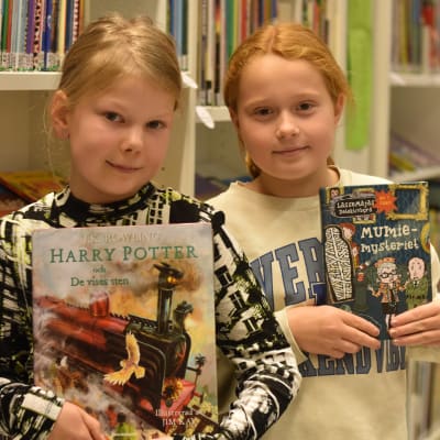 Två flickor står vid en hylla i ett bibliotek och håller i varsin favoritbok.