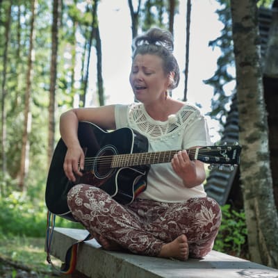 Muusikko Heidi Niinimäki soittaa kitaraa Alpo Jaakolan patsaspuistossa, ympärillä vihreää metsää.