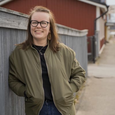 En kvinna står lutad mot ett staket och tittar in i kameran.