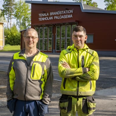 Två arbetsklädda (gula och gråsvarta kläder) män framför en ny brandstation. Tenala brandstation.