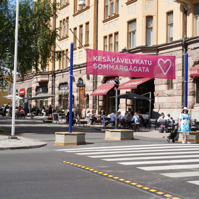 Ett stort övergångsställe framför en skylt där det står sommargågata. På gågatan sitter människor på uteserveringar.
