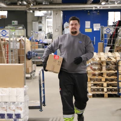Mathias Rosenlund går i en logistikcentral iklädd arbetskläder med ett paket under armen. 