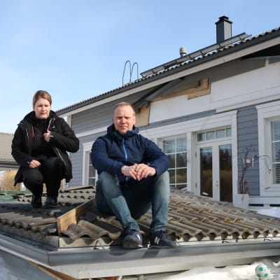 Mies ja nainen istuvat romahtaneen terassin katon päällä lumisella pihamaalla. Taustalla omakotitalo, jonka seinässä on reikä.