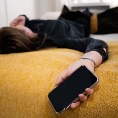 En person med långt hår ligger på en säng och håller i en telefon.