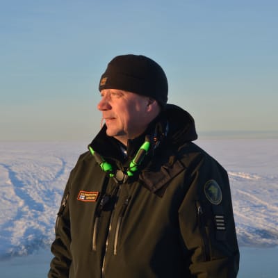 Man i sjöbevakningsform tittar förbi kameran. Han står framför snö- och istäckt hav.