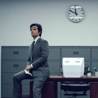 En allvarlig Mark S. (Adam Scott) med ett plåster på pannan, sitter på kanten av ett skrivbord i ett grått rum iklädd en grå kostym.