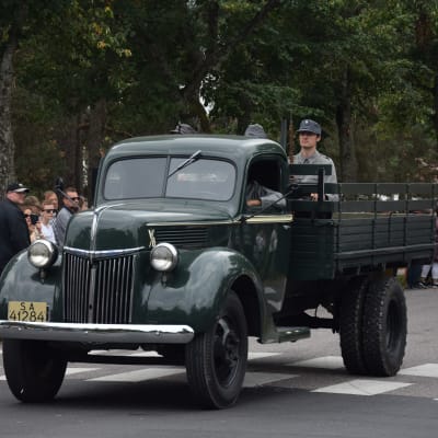 Militärfordon med tidsenligt klädda herrar i. Publik i bakgrunden.