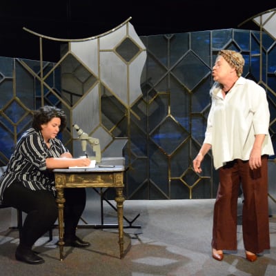Två kvinnor på teaterscen. En sitter och skriver i ett block, till höger står en annan kvinna. 