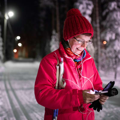 Keski-ikäinen nainen kuuntelee audiosisältöjä Yle Areenasta samalla kun on hiihtolenkillä talvisena iltana.