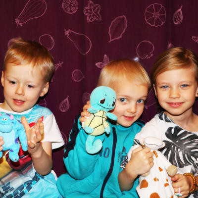 Tre barn, två pojkar till vänster och en flicka till höger, sitter på en bänk framför en lila gardin och håller varsitt kramdjur i handen.