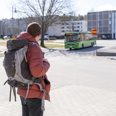 En man med röd vinterjacka, brun mössa och ryggsäck står med ryggen till och väntar på en grön buss som anländer.
