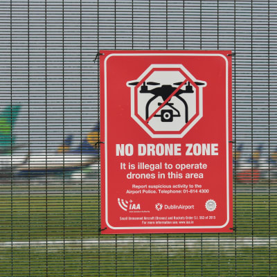 Droonin lennätyskieltokyltti aidassa lentokentän edessä.