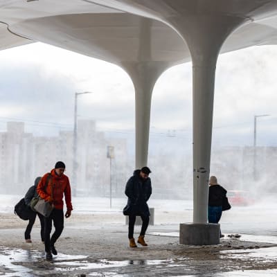 Ihmisiä kävelee Helsingin Pasilassa lumisateessa ja tuulessa.