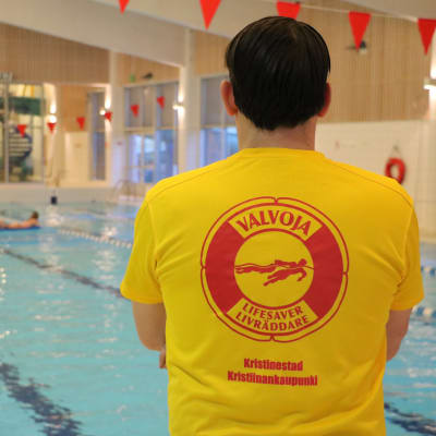 En simövervakare med gul tröja står bredvid en simbassäng.