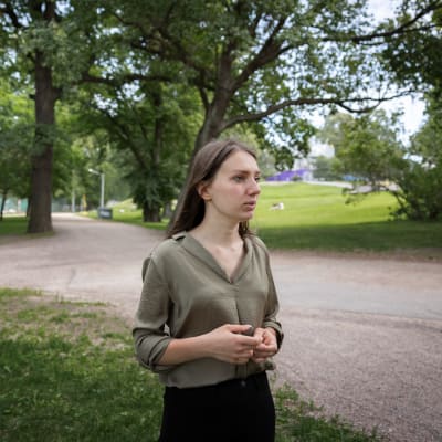 Vihreäpaitainen nainen seisoo puistossa katsoen kameran oikealle samalla puristaen sormiaan yhteen.
