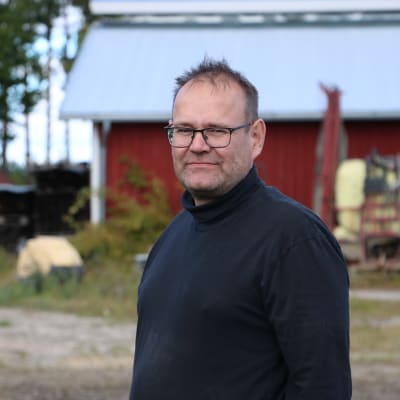 En man med kort hår och glasögon. I bakgrunden syns en röd lada.