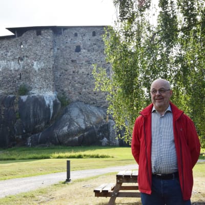 En man står i röd rock och skjorta. I bakgrunden syns en stenborg som är Raseborgs slottsruin. 