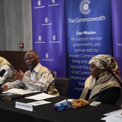 Fyra personer sitter bakom ett långt bord. Vid en plats finns det många mikrofoner. Där sitter Thabo Mbeki. Bakom honom syns Samväldets logotyp och texten The Commonwealth.