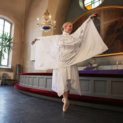 Kirkon alttarin edessä tanssii balettitossuissa nainen, jolla on valkoinen tanssiasu, jonka etumuksessa on risti.