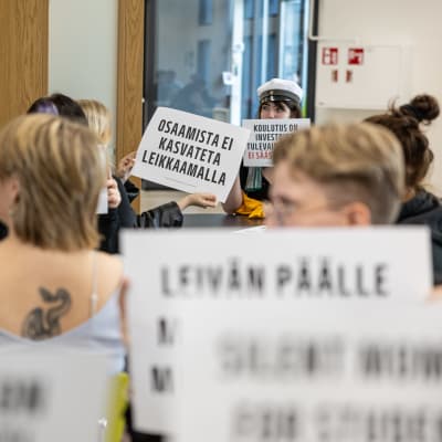 Opiskelijat istuvat pöytien ääressä ja pitävät iskulauseita sisältäviä kylttejä kädessään.