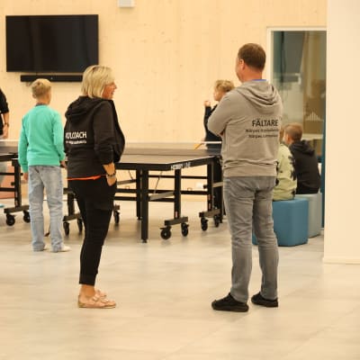 En kvinna och en man står och pratar med varandra. I bakgrunden syns ungdomar som spelar pingis.