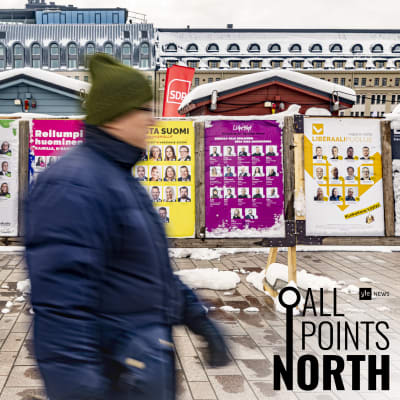 Sumeaksi jäävä henkilö kävelee vaalikylttien ohitse torilla. Kuvassa All Points North -logo.