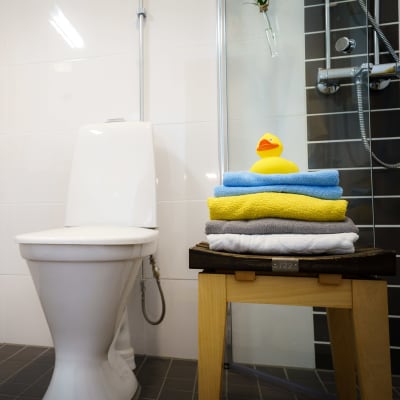 En wc-stol i ett kaklat badrum. Bredvid en pall med vikta handdukar och överst en gul badanka.