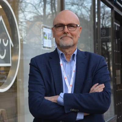 Mani 50-årsåldern tittar in i kameran. Han står utomhusoch har på sig en blå kavaj och blå skjorta. Han har glasögon.