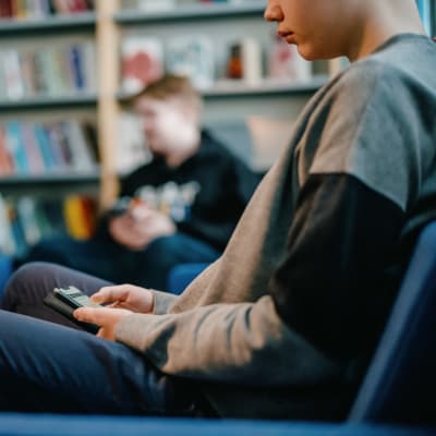 Tunnistamaton oppilas istuu sohvalla koulun kirjastossa käyttämässä puhelinta.