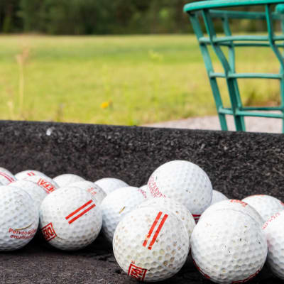 Flera golfbollar bredvid varandra på en grå yta. I bakgrunden syns gräs och en grön korg.