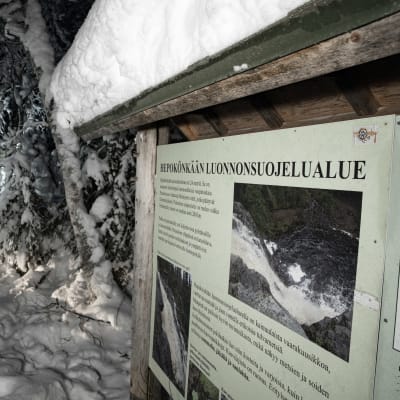 Hepokönkään luonnonsuojelualueen infotaulu lumisessa maisemassa ja taustalla hirsinen käymälä.