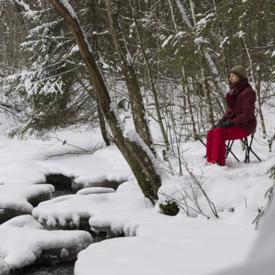 Nainen istuu jakkaralla puron varrella metsässä. 