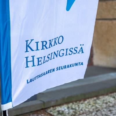 Lähikuva Kirkko Helsingissä -tekstistä Lauttasaaren seurakunnan sisäänkäynnin edustalla olevassa lipussa.