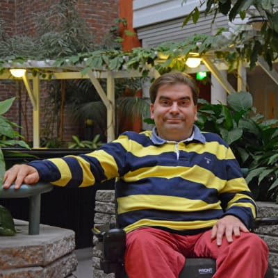En man sitter inomhus i en rullstol framför en massa gröna växter och träd. 