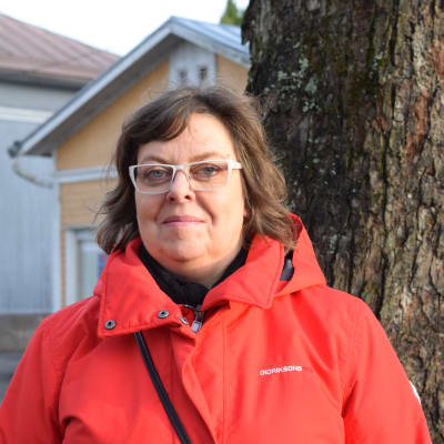 En kvinna med röd jacka och glasögon poserar ute vid ett större träd inne i en stad.