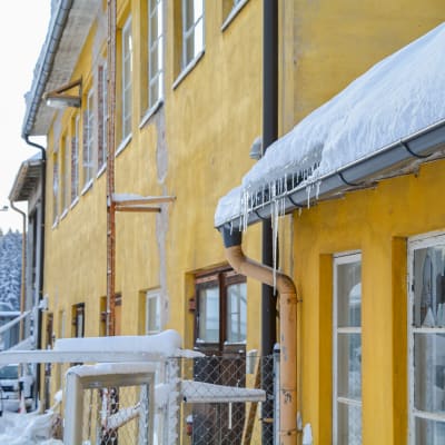 Närbild på gul gammal byggnad under vintertid. På taket ligger ett lager snö. 