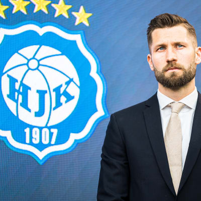Joona Toivio i klädd kostym och slips med HJK-logon i bakgrunden.