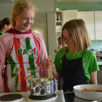 Två flickor står framför en kastrull full av pasta. Den ena har en ljusröd munkjacka och ett färggrant förkläde. Den andra har en grön t-skjorta och ett mörkt förkläde.