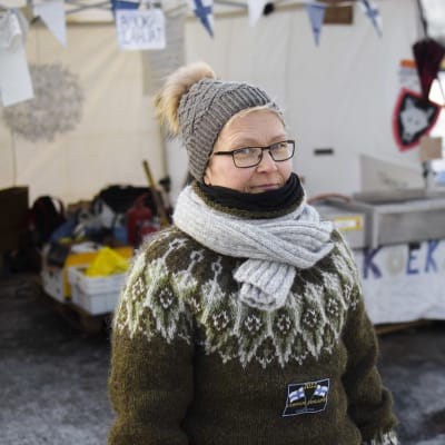 En kvinna står framför ett tält och har på sig varma kläder. Hon ser allvarlig ut och tittar in i kameran.