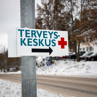 Tienvarteen kiinnitetty kyltti, jossa lukee "terveyskeskus". Taustalla näkyy luminen tie ja terveyskeskusrakennus.