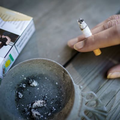 Naisen käsi pitelee savuketta pöydällä, jossa myös tuhkakuppi ja savukeaski.