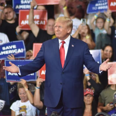På bilden syns ex-presidenten Donald Trump på ett av sina politiska evenemang. I bakgrunden står hans anhängare med skyltar i blått, rött och vitt där det står "Save America". 