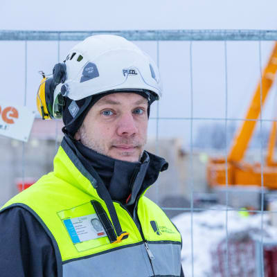 En man med skyddshjälm och väst tittar in i kameran. I bakgrunden ett stängsel och en lyftkran.